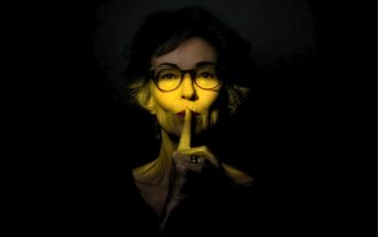 An older woman with glasses holds a finger to her lips in a “shush” gesture, with her face dramatically lit by a yellow spotlight against a dark background.
