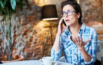A person in glasses and a checkered shirt is sitting at a table with a cup of coffee, talking on a phone with a concerned expression. The background features a brick wall, a lamp, and a plant.