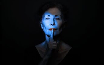 A woman with blue light on her face holds a finger to her lips, signaling silence. The background is dark, emphasizing her serious expression and the gesture.