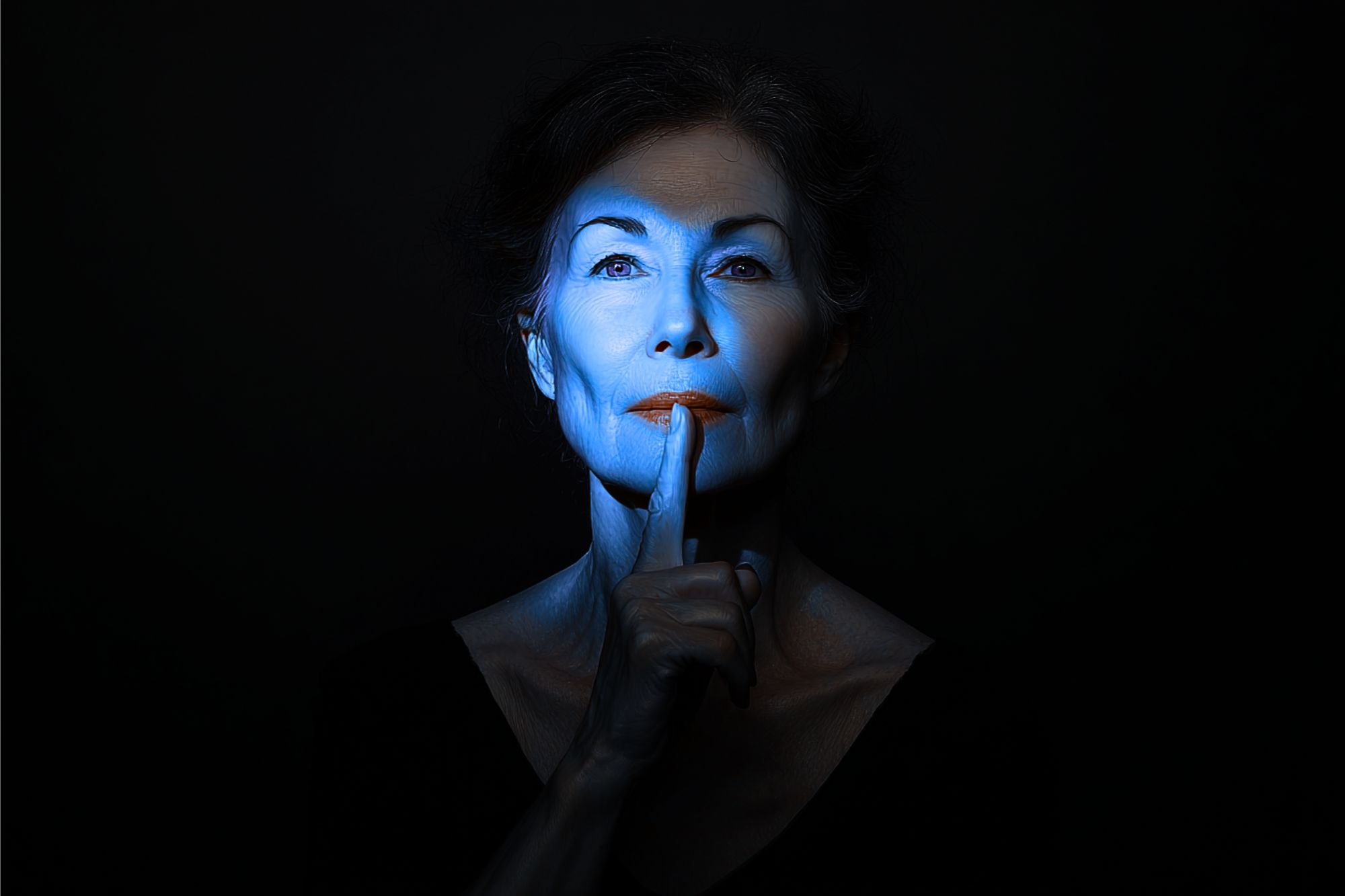 A woman with blue light on her face holds a finger to her lips, signaling silence. The background is dark, emphasizing her serious expression and the gesture.