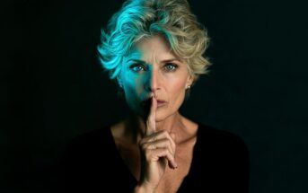 A woman with short, blonde hair holds a finger to her lips in a gesture for silence.