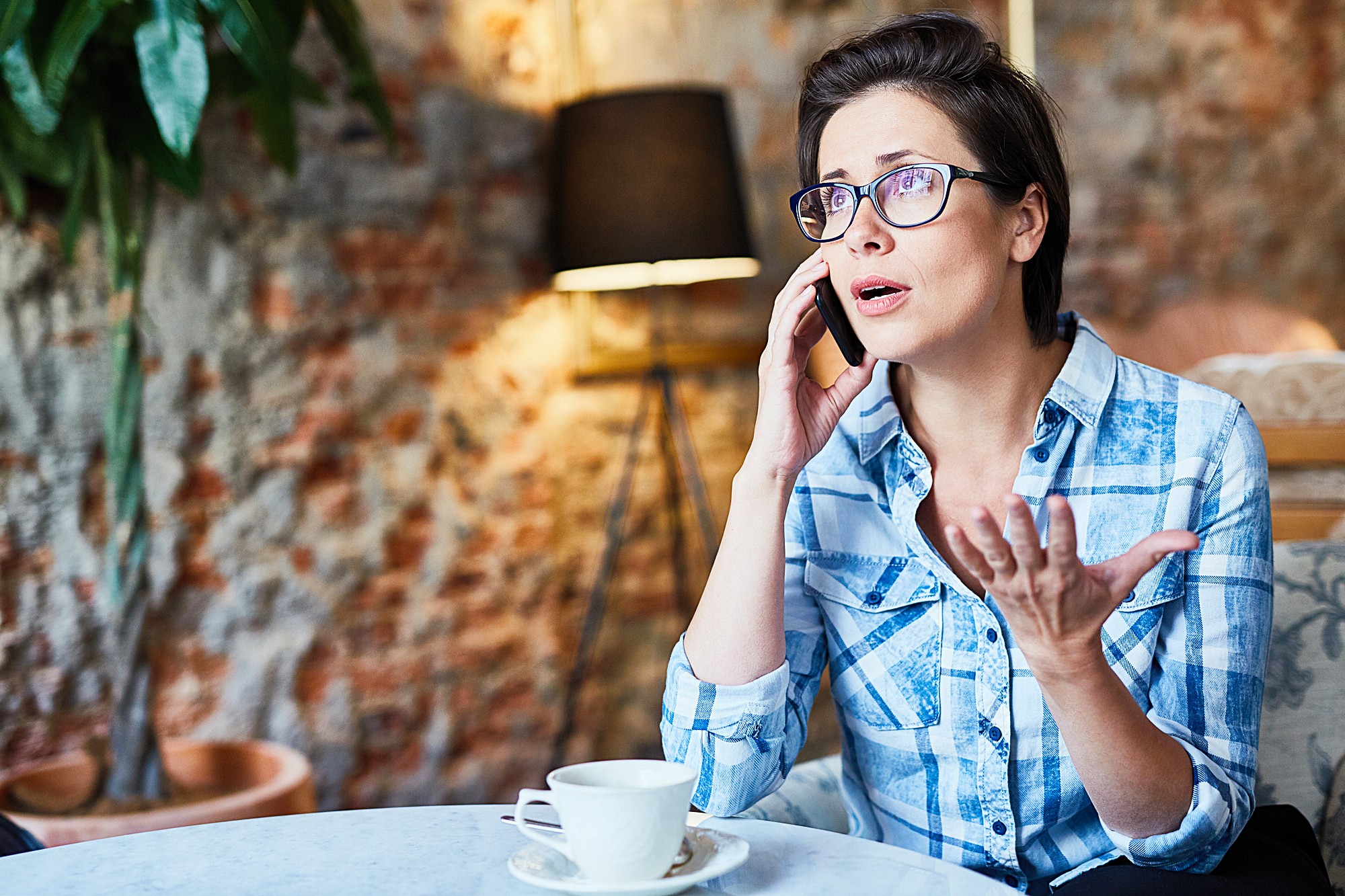 A person in glasses and a checkered shirt is sitting at a table with a cup of coffee, talking on a phone with a concerned expression. The background features a brick wall, a lamp, and a plant.