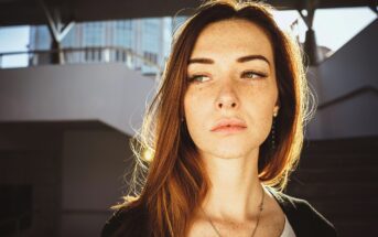 A young woman with long red hair and freckles stands in sunlight, looking to the side with a serious expression. The background shows a modern, partially shaded outdoor structure.