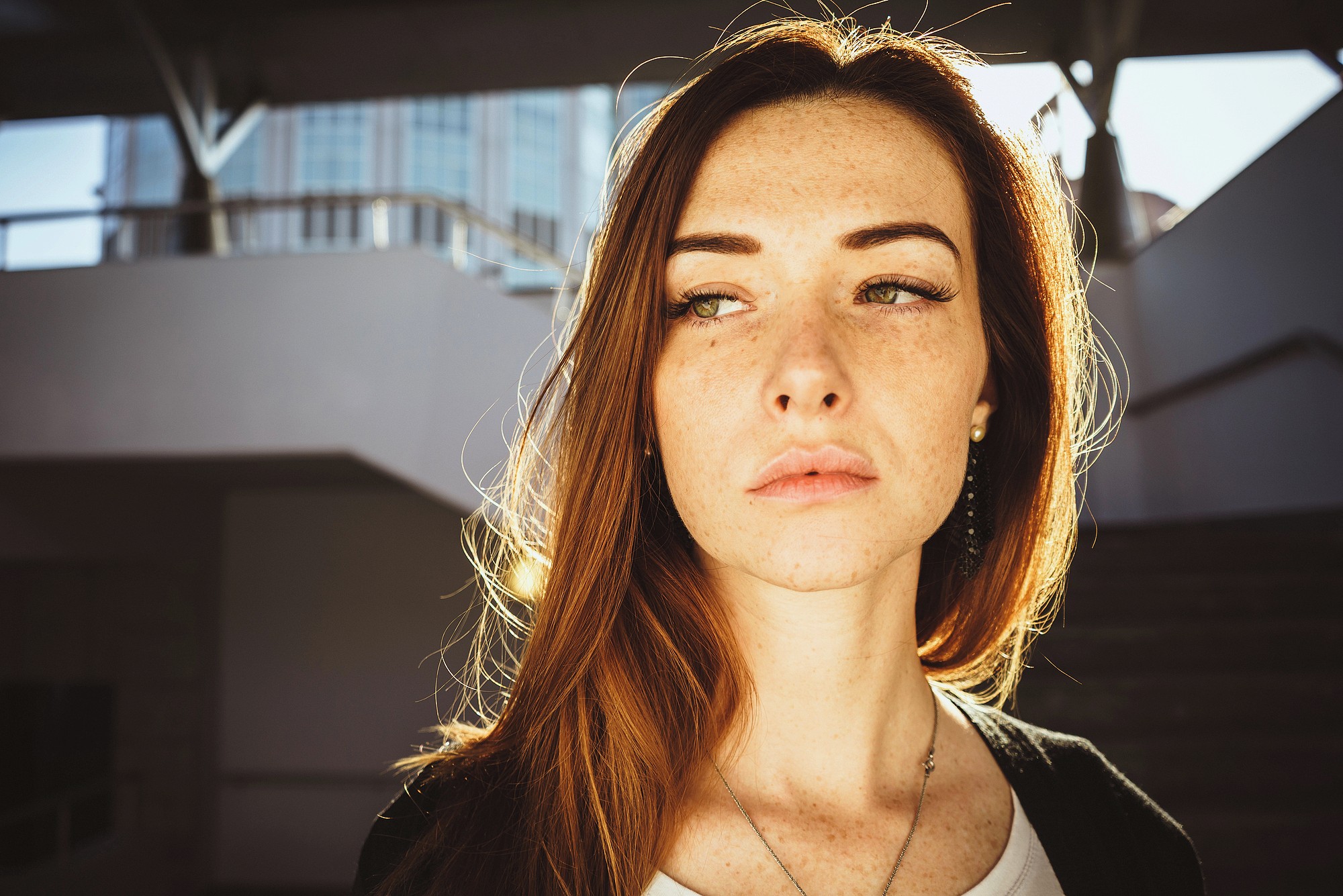 A young woman with long red hair and freckles stands in sunlight, looking to the side with a serious expression. The background shows a modern, partially shaded outdoor structure.