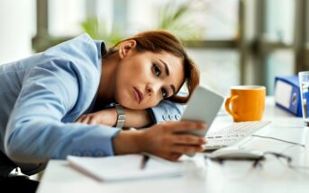 A woman in a blue blazer rests her head on her arms at a desk, looking tired while holding a smartphone. A keyboard, notebook, orange mug, and eyeglasses are on the desk in front of her.