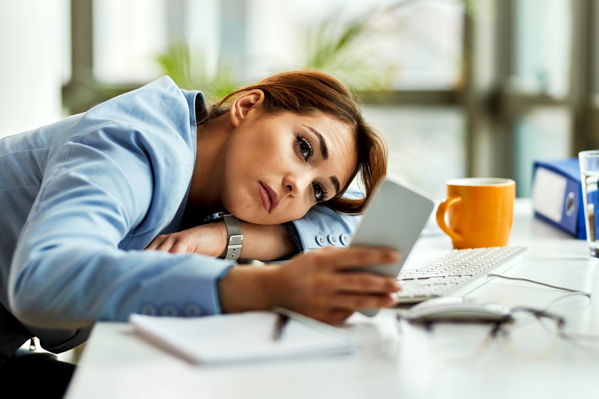 A woman in a blue blazer rests her head on her arms at a desk, looking tired while holding a smartphone. A keyboard, notebook, orange mug, and eyeglasses are on the desk in front of her.