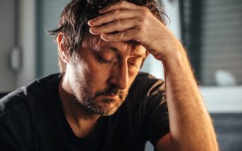 A man with tousled hair rests his forehead on his hand, appearing deep in thought or concern. He wears a dark t-shirt, and the background is softly blurred, emphasizing his expression. The lighting is warm and moody.