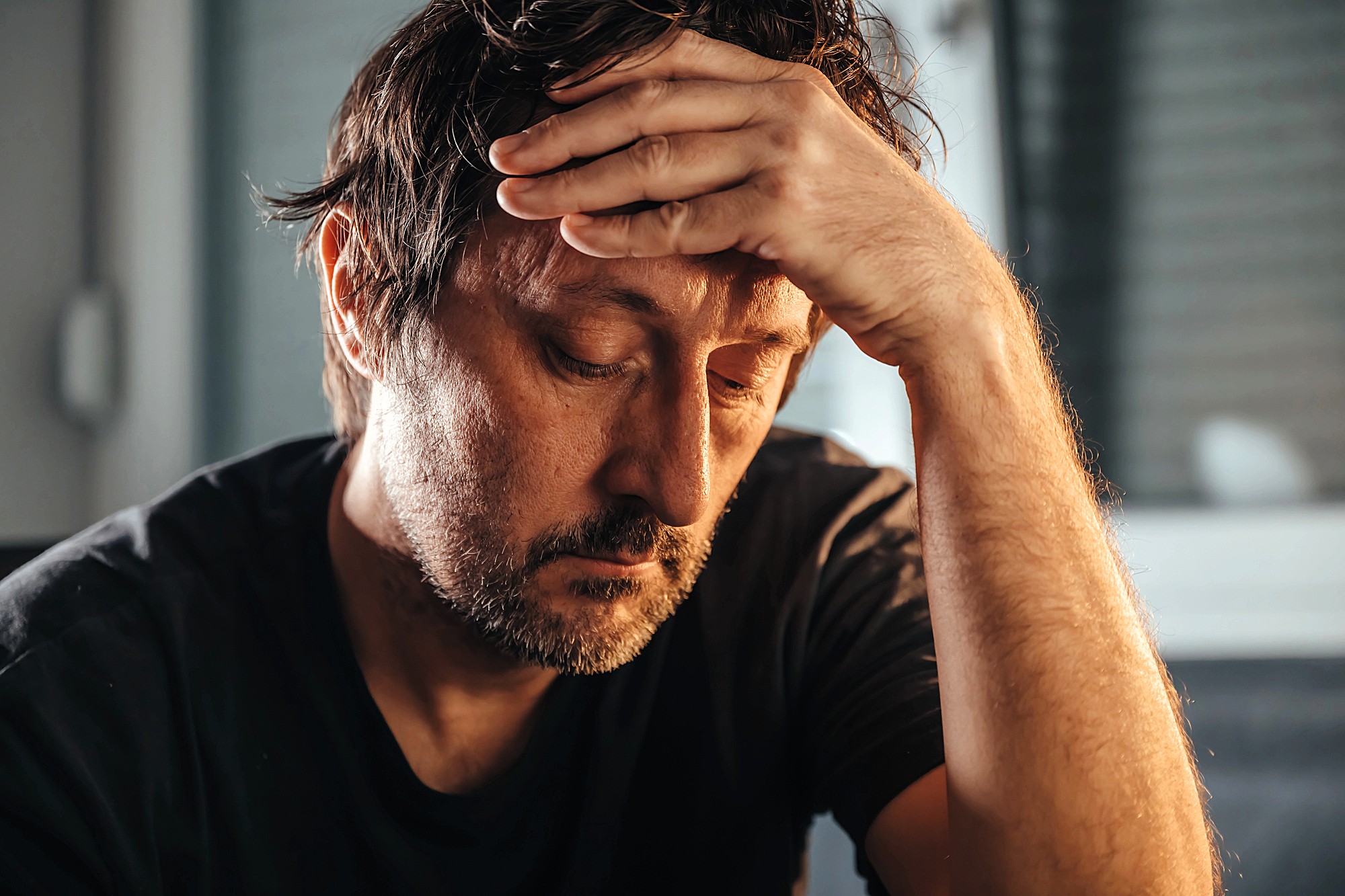 A man with tousled hair rests his forehead on his hand, appearing deep in thought or concern. He wears a dark t-shirt, and the background is softly blurred, emphasizing his expression. The lighting is warm and moody.