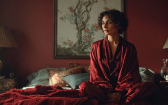 A woman with curly hair, wearing a deep red robe, sits cross-legged on a bed with red bedding in a warmly lit room. Behind her is a large painting of a tree with flowers on a red wall.