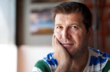A man with short dark hair and stubble rests his chin on his hand, looking at the camera. He wears a striped shirt in shades of blue, green, and white. The background is softly blurred, highlighting his thoughtful expression.