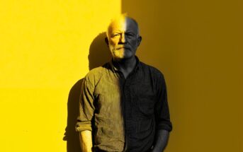 An older man with a beard stands against a bright yellow wall. Half of his face and body are in shadow, creating a stark contrast between light and dark. He has his hands in his pockets and looks directly at the camera.