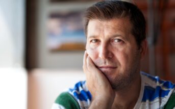 A man with short dark hair and stubble rests his chin on his hand, looking at the camera. He wears a striped shirt in shades of blue, green, and white. The background is softly blurred, highlighting his thoughtful expression.