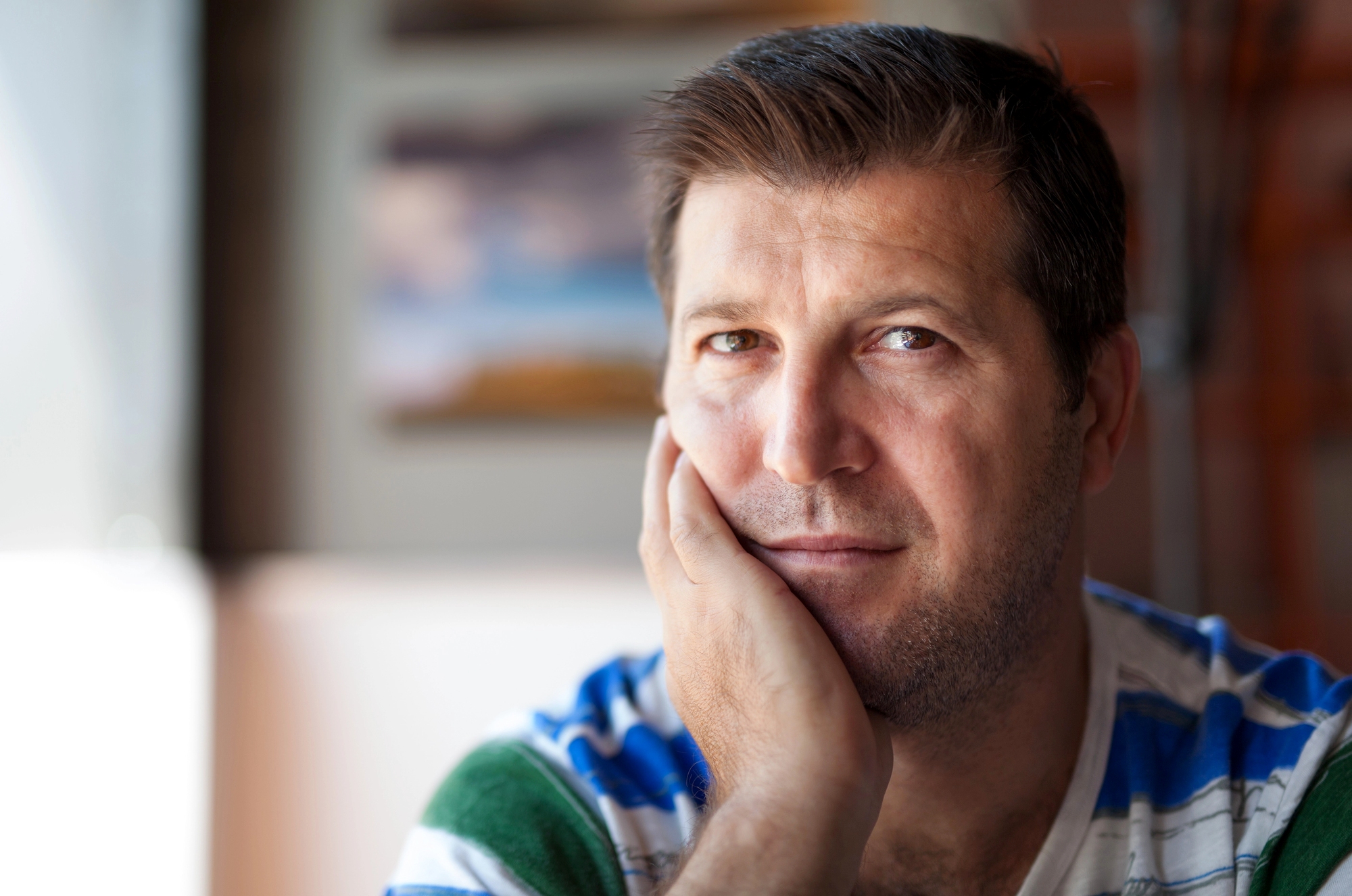 A man with short dark hair and stubble rests his chin on his hand, looking at the camera. He wears a striped shirt in shades of blue, green, and white. The background is softly blurred, highlighting his thoughtful expression.