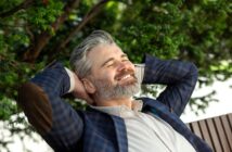 A smiling man with gray hair and a beard leans back with his hands behind his head. He is wearing a plaid blazer with elbow patches and a light-colored shirt. Green foliage is visible in the background, suggesting a relaxed setting.