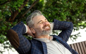 A smiling man with gray hair and a beard leans back with his hands behind his head. He is wearing a plaid blazer with elbow patches and a light-colored shirt. Green foliage is visible in the background, suggesting a relaxed setting.