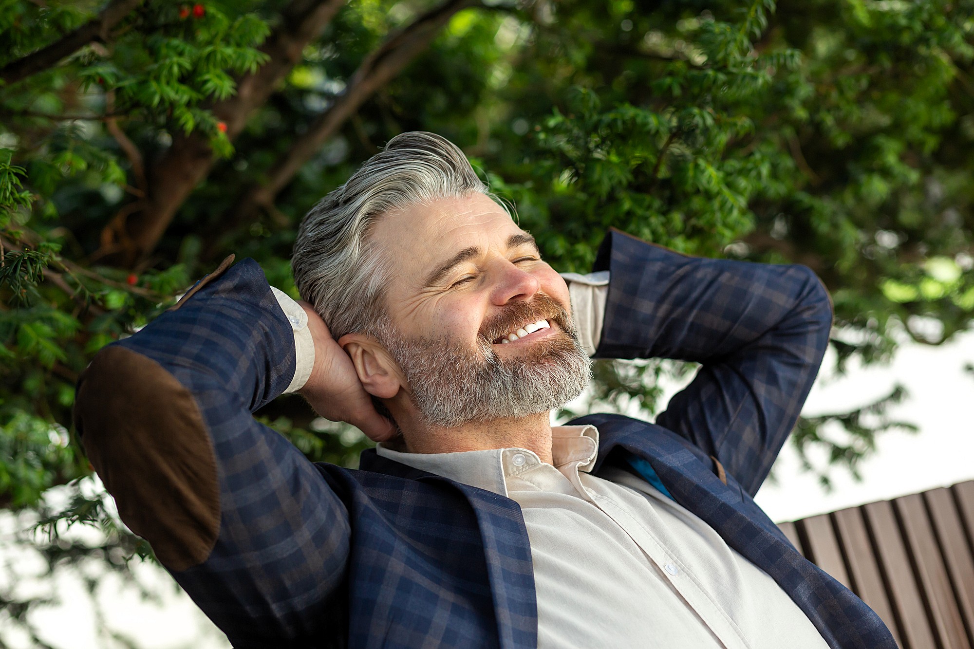 A smiling man with gray hair and a beard leans back with his hands behind his head. He is wearing a plaid blazer with elbow patches and a light-colored shirt. Green foliage is visible in the background, suggesting a relaxed setting.