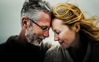 An older man and woman gently touch foreheads, eyes closed and smiling, with golden light sparkles between them. The background is softly blurred, and the woman’s hair flows in the wind.