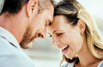 A happy man and woman stand close together, touching their foreheads and smiling warmly. The woman laughs with her eyes closed, and both appear joyful and affectionate in a sunlit outdoor setting.