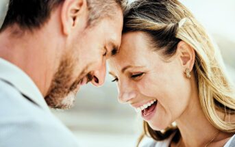 A happy man and woman stand close together, touching their foreheads and smiling warmly. The woman laughs with her eyes closed, and both appear joyful and affectionate in a sunlit outdoor setting.