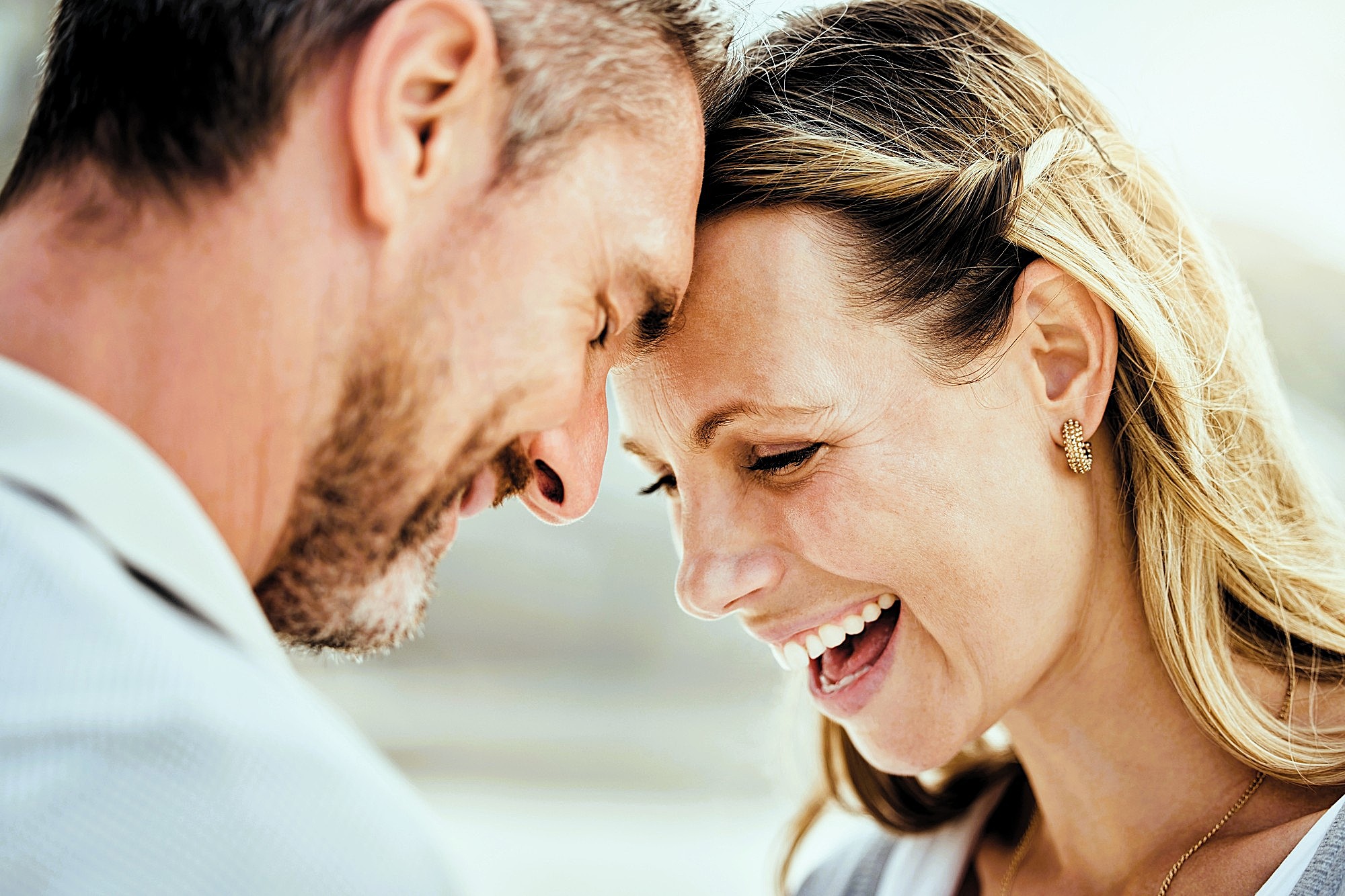 A happy man and woman stand close together, touching their foreheads and smiling warmly. The woman laughs with her eyes closed, and both appear joyful and affectionate in a sunlit outdoor setting.