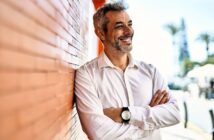 A man with gray hair and a beard, wearing a white shirt and a black watch, stands smiling with arms crossed against an orange brick wall. The background is blurred, showing trees and a bright sky.
