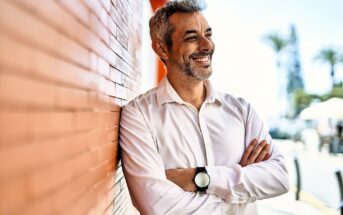 A man with gray hair and a beard, wearing a white shirt and a black watch, stands smiling with arms crossed against an orange brick wall. The background is blurred, showing trees and a bright sky.