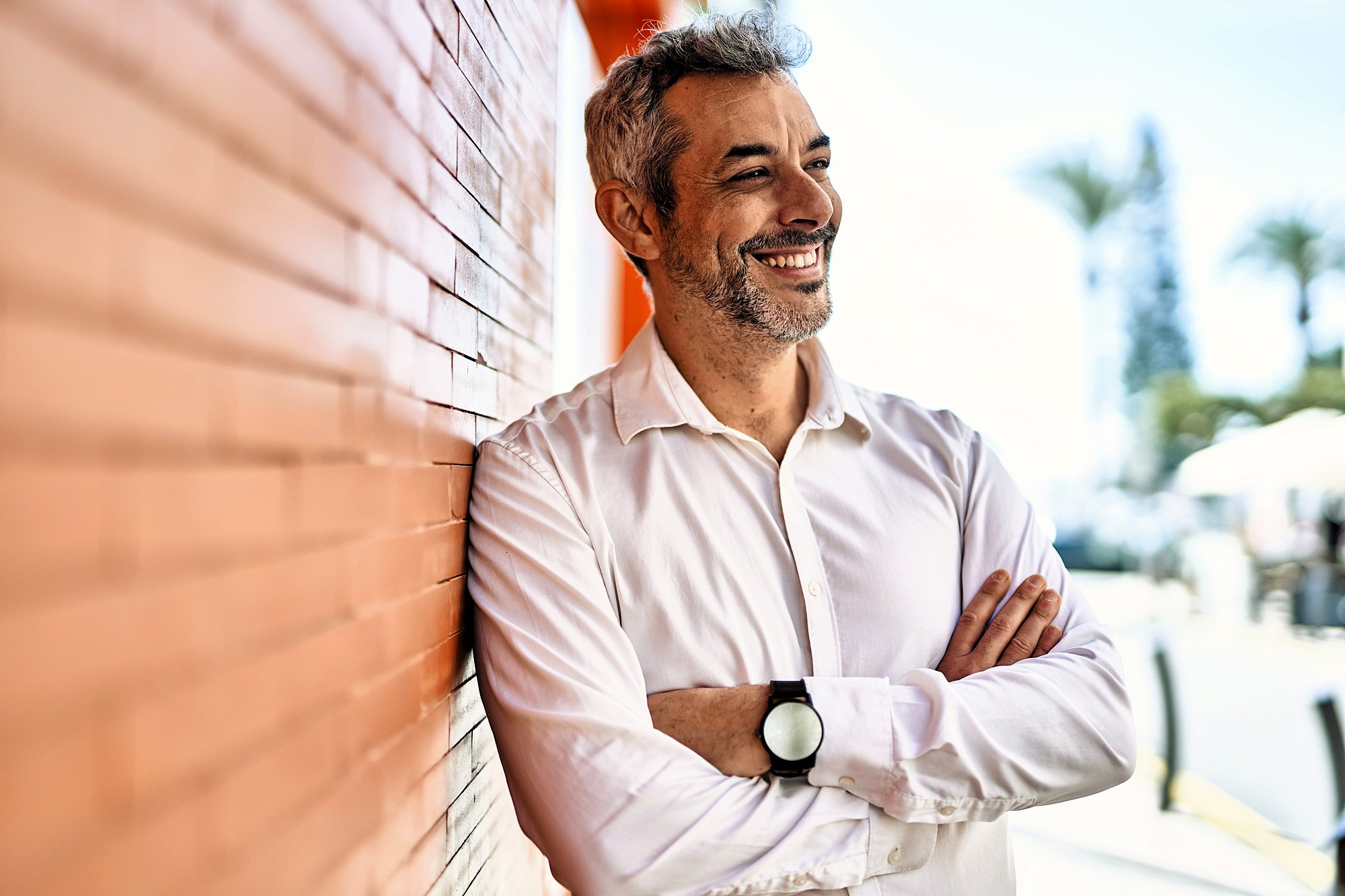 A man with gray hair and a beard, wearing a white shirt and a black watch, stands smiling with arms crossed against an orange brick wall. The background is blurred, showing trees and a bright sky.