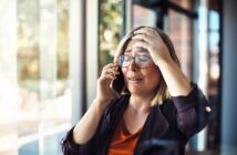 A woman with short blonde hair and glasses is standing by a window, holding a phone to her ear with one hand and touching her forehead with the other. She appears distressed, with her eyes closed. Sunlight streams in through the window behind her.