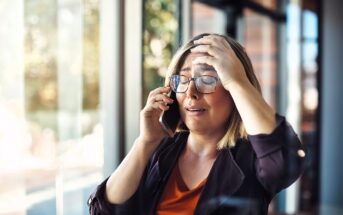 A woman with short blonde hair and glasses is standing by a window, holding a phone to her ear with one hand and touching her forehead with the other. She appears distressed, with her eyes closed. Sunlight streams in through the window behind her.