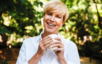 A smiling person with short blonde hair holds a white mug, standing outdoors amidst green foliage. They are wearing a white shirt and appear happy and relaxed in this natural setting.