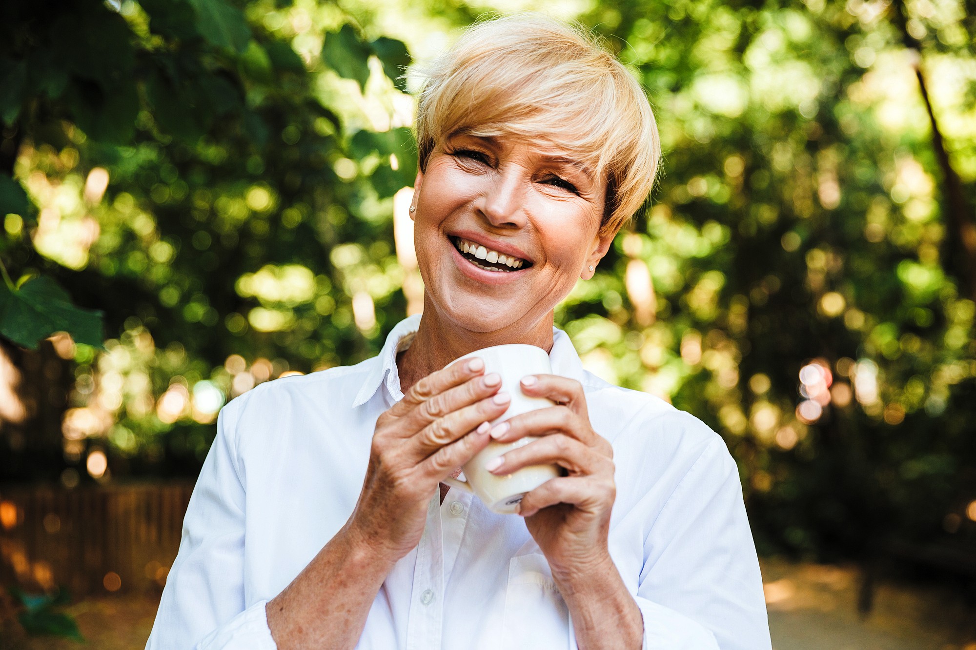 A smiling person with short blonde hair holds a white mug, standing outdoors amidst green foliage. They are wearing a white shirt and appear happy and relaxed in this natural setting.