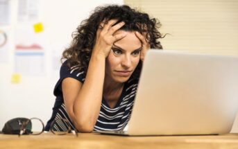 A woman with curly hair sits at a desk, looking stressed and holding her head with both hands while staring at a laptop screen. Glasses and papers are on the desk beside her.