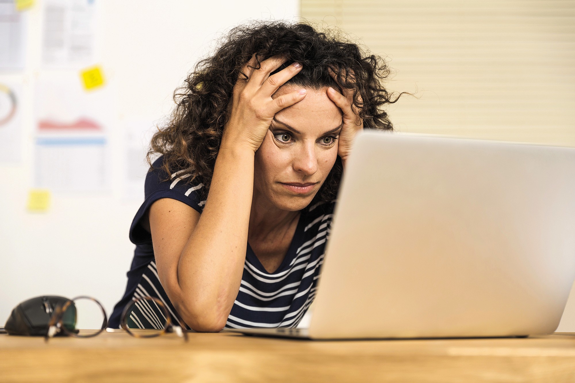 A woman with curly hair sits at a desk, looking stressed and holding her head with both hands while staring at a laptop screen. Glasses and papers are on the desk beside her.