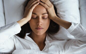 A woman lies in bed with her eyes closed, holding her hands to her forehead, appearing stressed or unwell. She is wearing a white long-sleeve shirt and is partially covered by a white blanket.