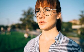 A young woman with red hair and round glasses stands outside in sunlight, wearing a blue and white striped shirt, looking ahead with a serious expression. The background is blurred greenery and trees.