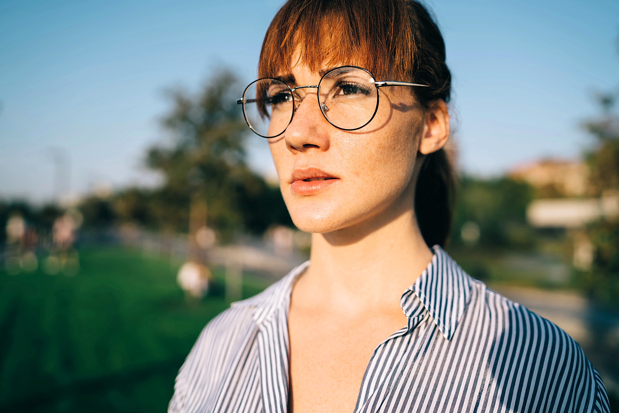 A young woman with red hair and round glasses stands outside in sunlight, wearing a blue and white striped shirt, looking ahead with a serious expression. The background is blurred greenery and trees.