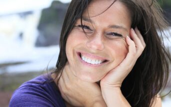 A woman with long brown hair smiles brightly while resting her head on her hand. She is wearing a purple top and is outdoors, with a blurred natural background behind her.