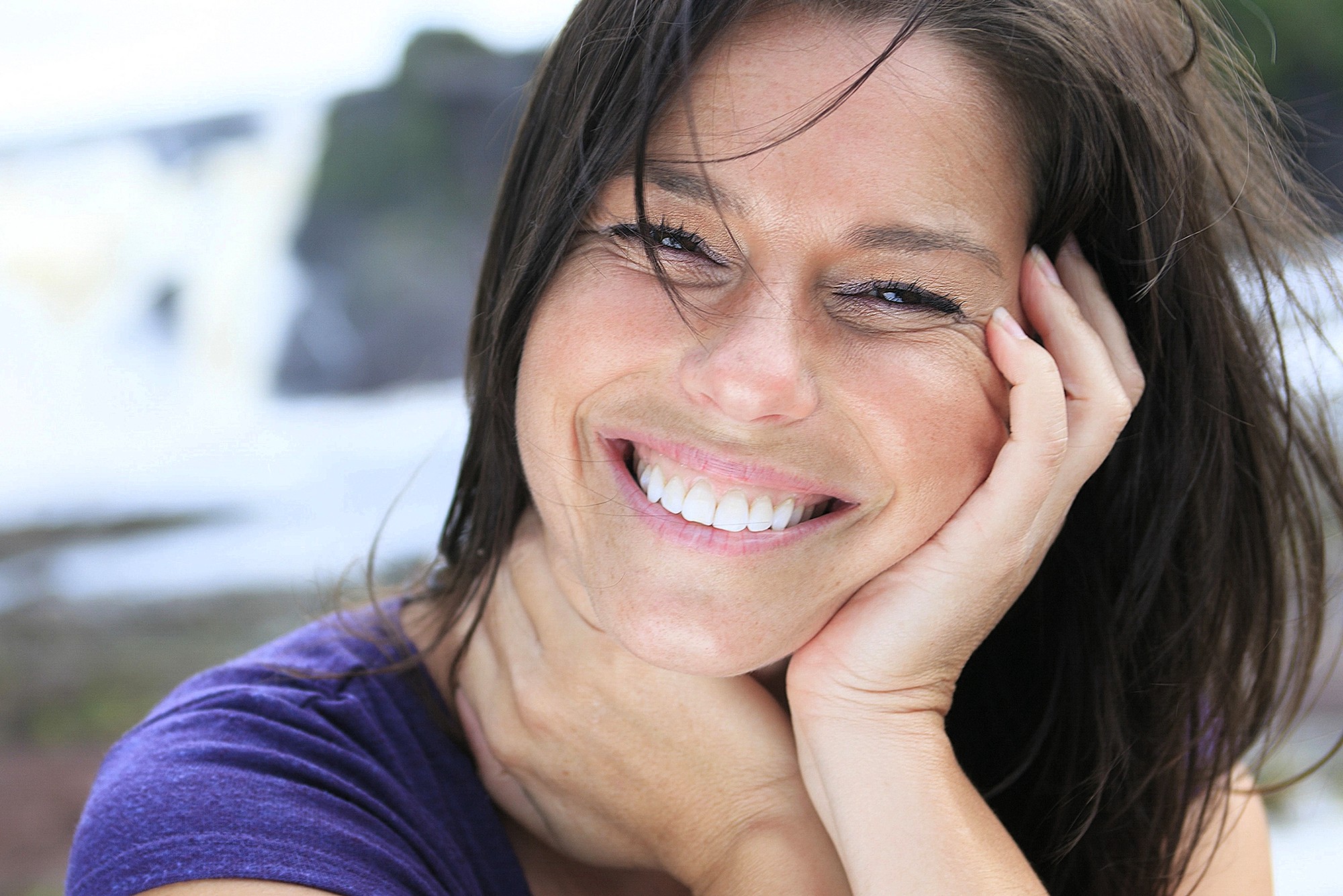 A woman with long brown hair smiles brightly while resting her head on her hand. She is wearing a purple top and is outdoors, with a blurred natural background behind her.