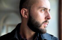 A man with a short beard and closely trimmed hair looks thoughtfully out of a window. He is wearing a black jacket and a dark shirt with small white dots. The lighting highlights his face and profile.