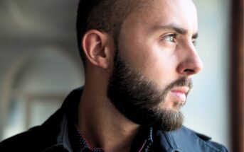 A man with a short beard and closely trimmed hair looks thoughtfully out of a window. He is wearing a black jacket and a dark shirt with small white dots. The lighting highlights his face and profile.