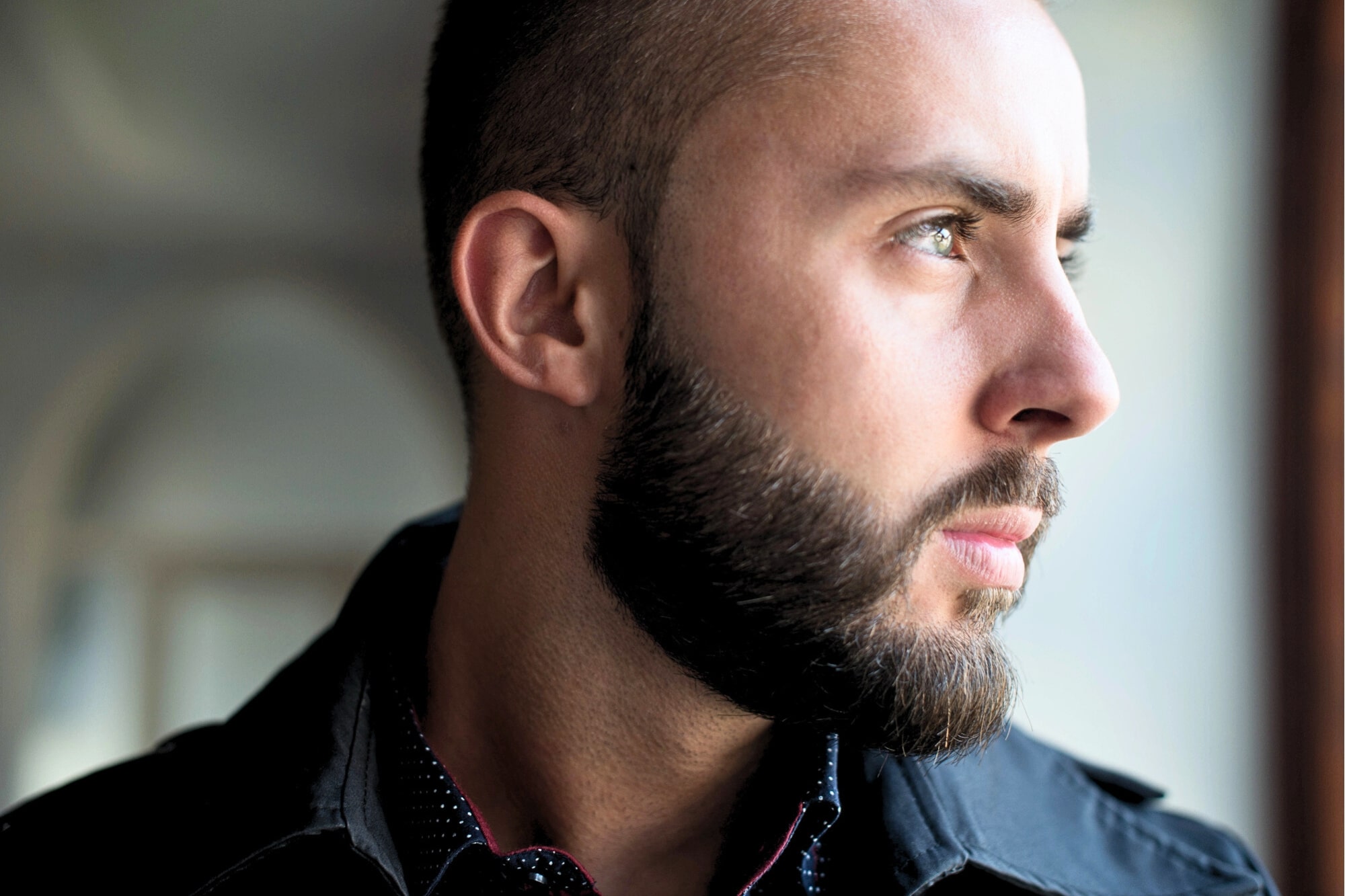 A man with a short beard and closely trimmed hair looks thoughtfully out of a window. He is wearing a black jacket and a dark shirt with small white dots. The lighting highlights his face and profile.