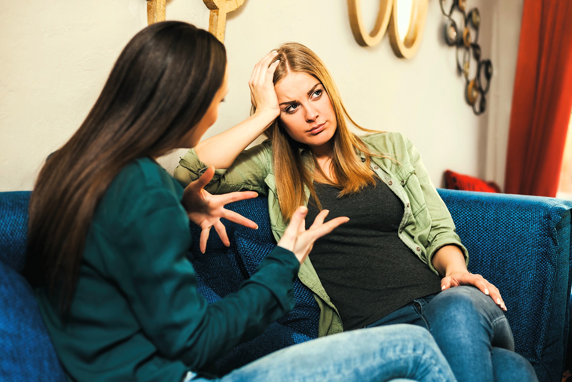 Two women sit on a blue couch. One, with long brown hair, gestures expressively while talking. The other, with blonde hair, rests her head on her hand, appearing thoughtful or concerned. The background includes wall decor and a red curtain.