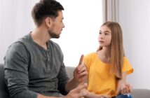 A man and a girl are sitting on a couch having a conversation. The man gestures with his hand while speaking, and the girl listens attentively, wearing a yellow shirt. They appear to be in a bright room with curtains and a soft couch.