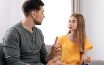 A man and a girl are sitting on a couch having a conversation. The man gestures with his hand while speaking, and the girl listens attentively, wearing a yellow shirt. They appear to be in a bright room with curtains and a soft couch.