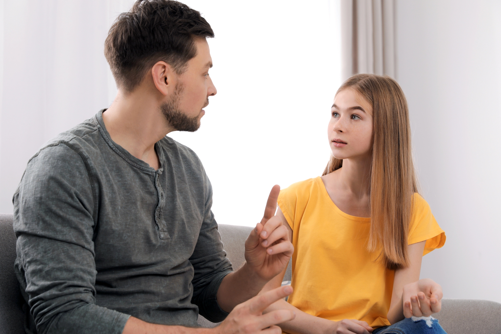 A man and a girl are sitting on a couch having a conversation. The man gestures with his hand while speaking, and the girl listens attentively, wearing a yellow shirt. They appear to be in a bright room with curtains and a soft couch.
