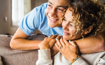 A young person in a blue shirt hugs a smiling adult from behind on a couch. The adult has curly hair and wears a light-colored sweater with colorful beaded bracelets. They both look happy and relaxed.