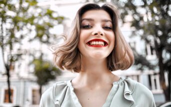 A young woman with short, wavy blonde hair and bright red lipstick smiles broadly outdoors, wearing a light green blouse. Trees and blurred buildings are visible in the background.