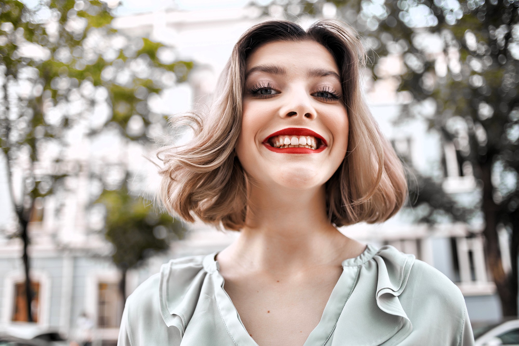 A young woman with short, wavy blonde hair and bright red lipstick smiles broadly outdoors, wearing a light green blouse. Trees and blurred buildings are visible in the background.