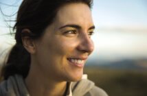 A person with long dark hair smiles while gazing to the side. The background is a blurred natural setting, suggesting an outdoor location. The lighting is warm and soft, casting a gentle glow on the person's face.