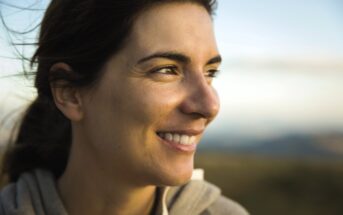 A person with long dark hair smiles while gazing to the side. The background is a blurred natural setting, suggesting an outdoor location. The lighting is warm and soft, casting a gentle glow on the person's face.